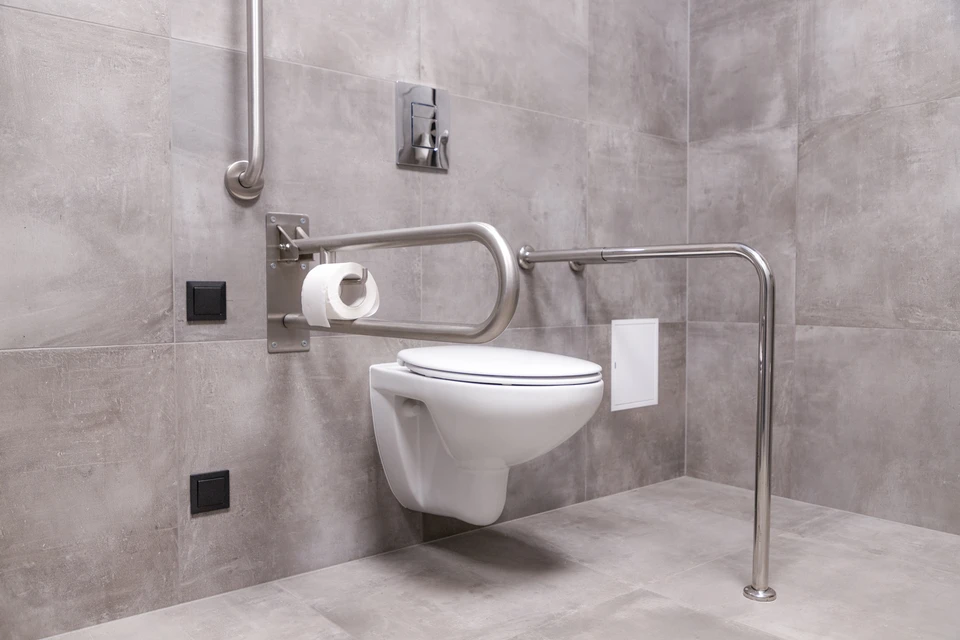 Salle de bains modeste en cours de rénovation : douche à l'italienne de plain-pied en carrelage gris antidérapant finition mate, deux barres d'appui chromées 35 cm installées verticalement sur carrelage blanc, bac de douche encastré, paroi vitrée fixe. Focus sur les barres d'appui mural montées sur carrelage et le sol antidérapant, lumière naturelle douce.