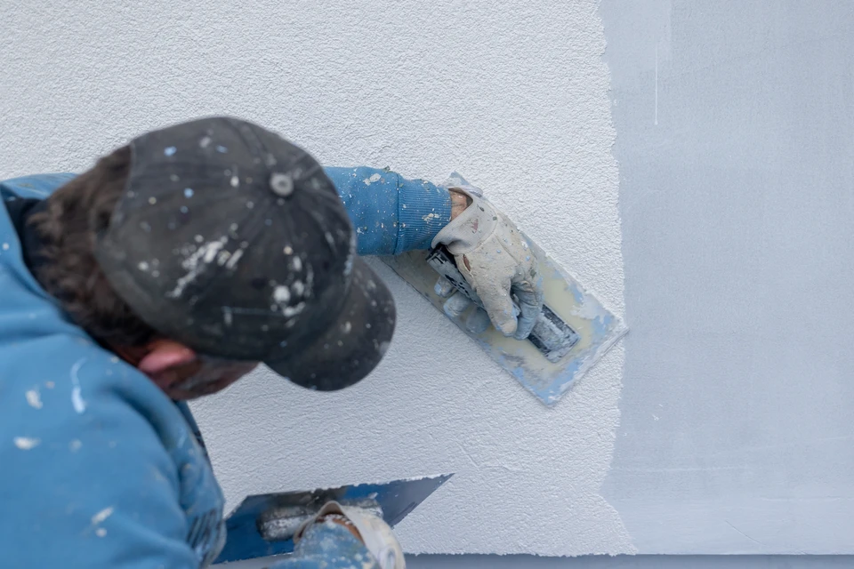Équipe de trois artisans professionnels sur un chantier de rénovation intérieure, en tenue de travail appropriée, posant ensemble devant un mur en cours de peinture ou d'enduit. L'un d'eux tient un outil métier (rouleau à peinture ou taloche). Arrière-plan : murs partiellement rénovés, bâche de protection au sol, contexte authentique de chantier résidentiel. Composition naturelle et professionnelle, lumière claire, ambiance de travail en équipe.